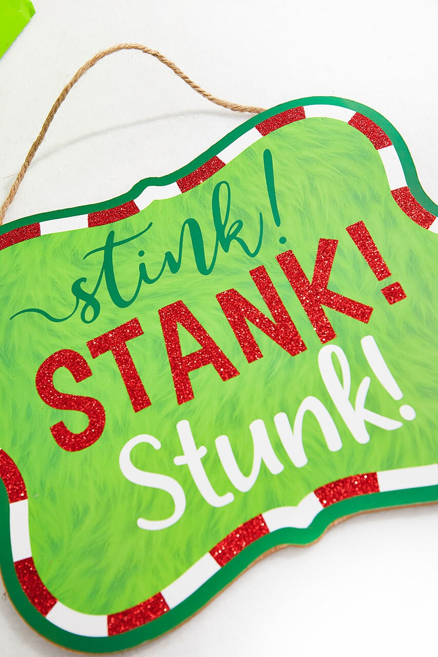 Hanging holiday sign that reads "stink! STANK! Stunk!" in a green, red glitter and white font. The background is designed to look like green fur. There's a red and white border up against a solid green border. Hangs from a jute rope.