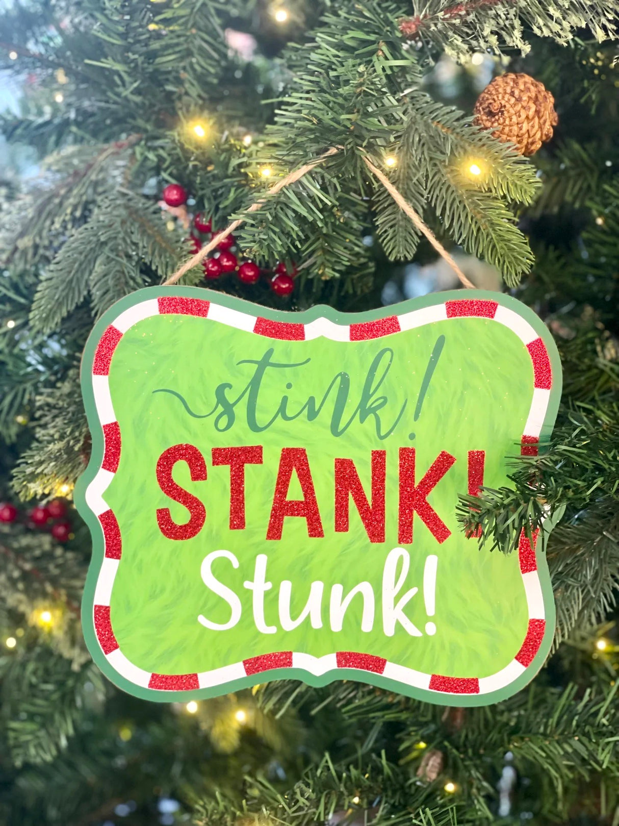 Hanging holiday sign that reads "stink! STANK! Stunk!" in a green, red glitter and white font. The background is designed to look like green fur. There's a red and white border up against a solid green border. Hangs from a jute rope.