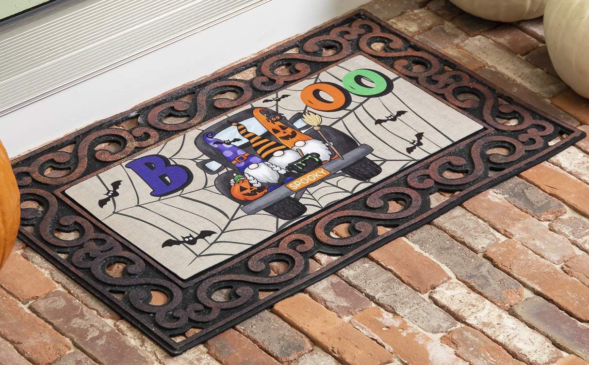 Doormat for Halloween. Reads "BOO" and features three gnomes in the back of a truck. One is holding a pumpkin, one is holding a cauldron and one is holding a broomstick. The license plate reads "SPOOKY" and there are bats and spiderweb accents.