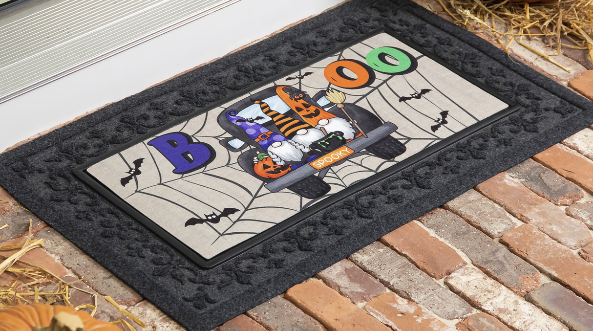 Doormat for Halloween. Reads "BOO" and features three gnomes in the back of a truck. One is holding a pumpkin, one is holding a cauldron and one is holding a broomstick. The license plate reads "SPOOKY" and there are bats and spiderweb accents.