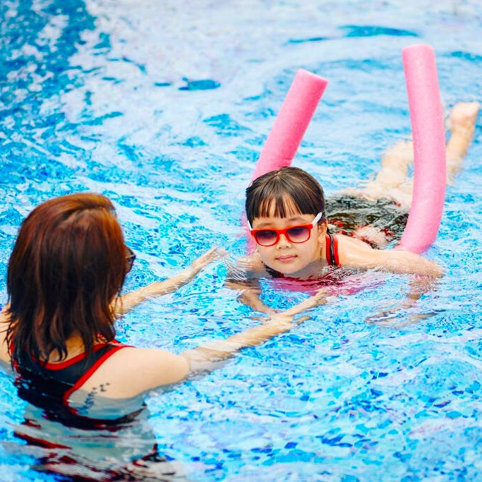 This is a lifestyle image showing a young girl learning to swim using a pink noodle while her mom helps.