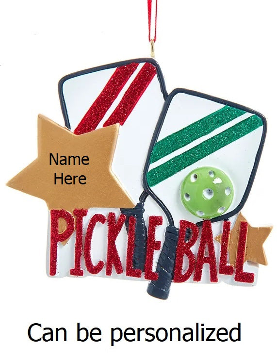 Ornament shaped like two white paddles with festive red and green stripes, as well as two gold stars. It reads "Pickleball" in red glitter.