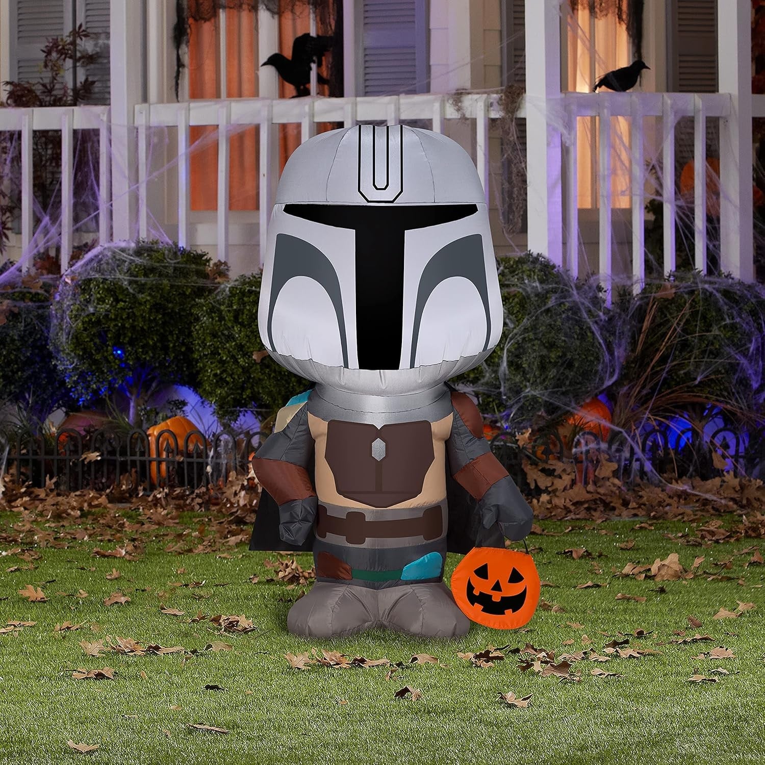 Halloween inflatable shaped like The Mandalorian from the Star Wars universe. He's holding a treat tote bag that looks like a carved pumpkin.