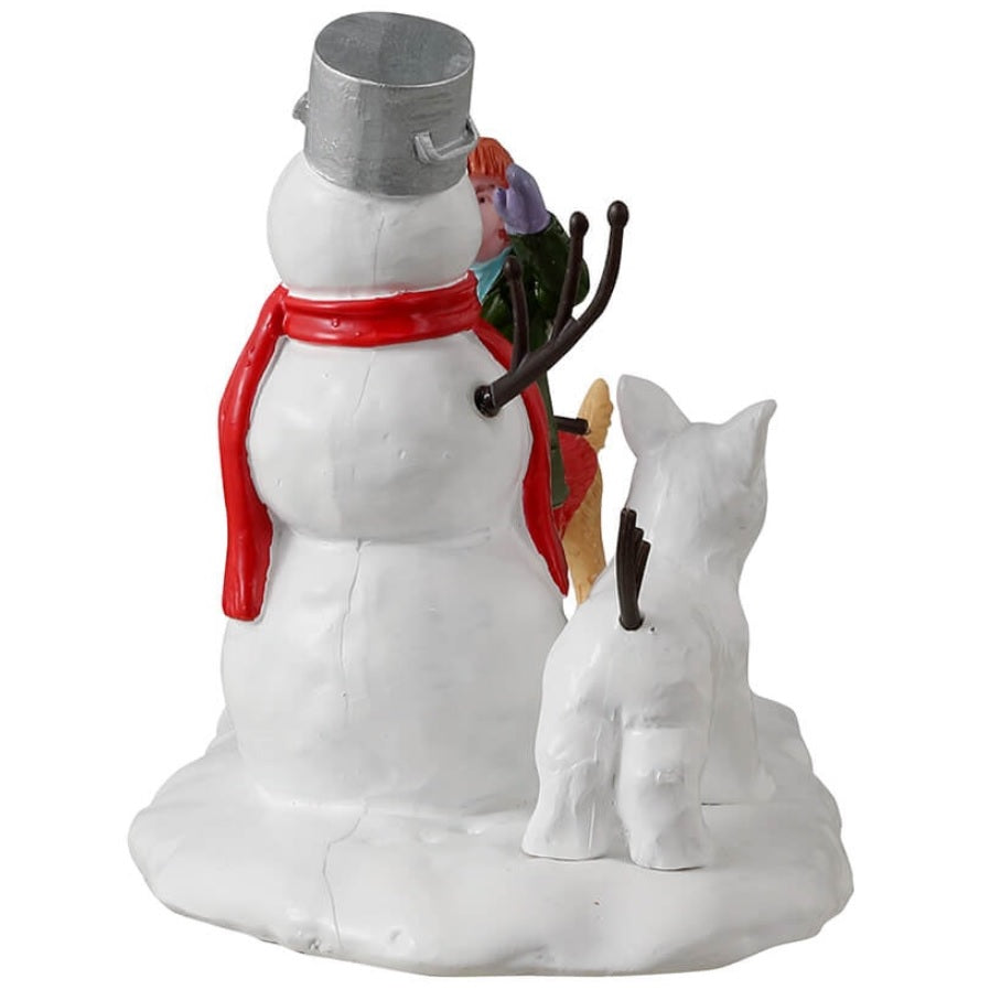 Resin village accent for a Lemax display. Designed to look like a young girl putting the finishing touches on a snowman. There's a dog holding a twig, trying to help. There's a dog made out of snow standing next to the snowman.