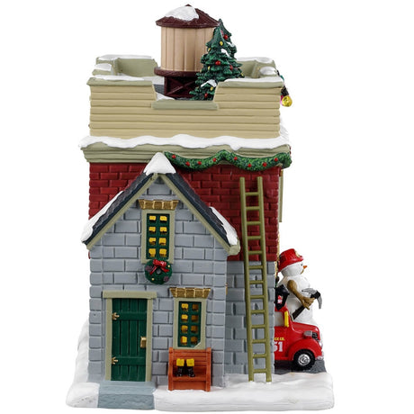 Porcelain lighted building for a Lemax display. Designed to look like a fire house decorated for Christmas. There are decorated trees and multi-colored lights on the roof. There are ladders on both sides of the building. In the window, you can see a decorated tree inside. There's a red firetruck parked in the garage and a snowman on the sidewalk.