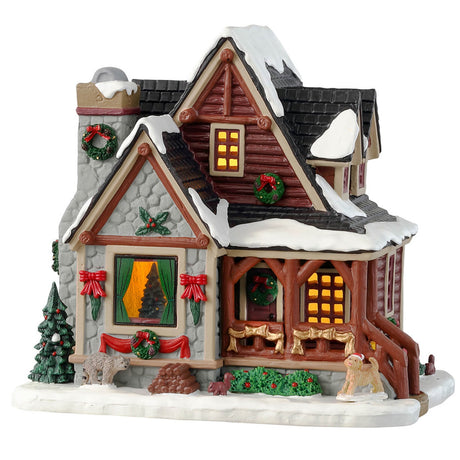 Porcelain lighted building for a Lemax display. Designed to look like a snow-covered log cabin. There are decorated wreaths, garland and Christmas trees. There's also a dog wearing a Santa hat near the front porch.