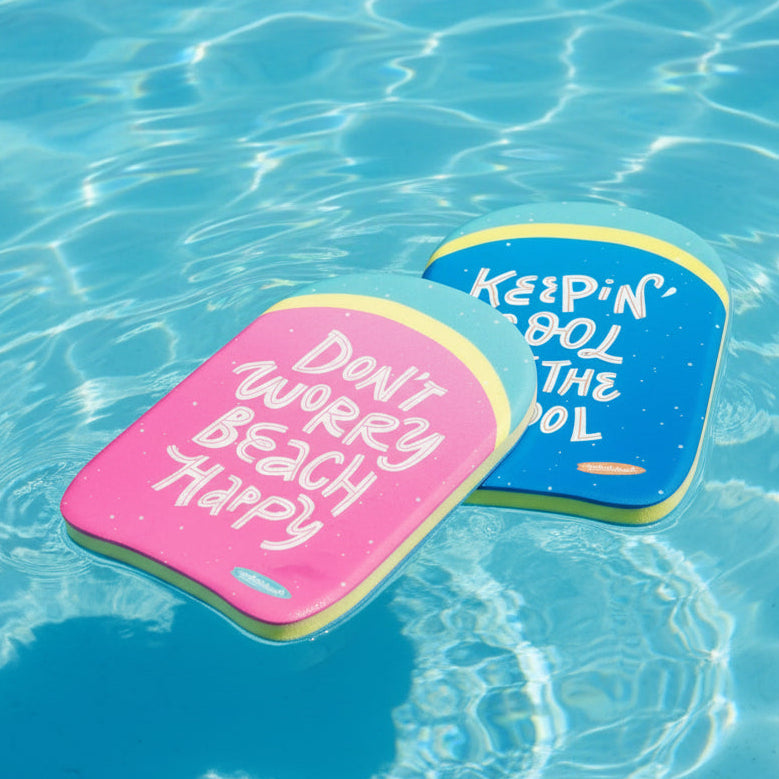 This image shows two kickboards resting on top of the water. One is pink and reads "Don't Worry Beach Happy" and the other is blue and reads "Keepin' Cool in the Pool."