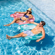 This is a lifestyle image showing two people in a swimming pool sitting in two different floating pool chairs. One is pink and one is blue.