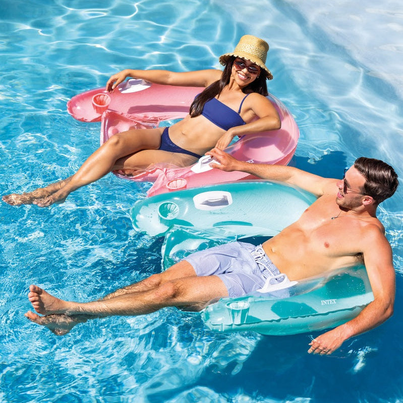 This is a lifestyle image showing two people in a swimming pool sitting in two different floating pool chairs. One is pink and one is blue.
