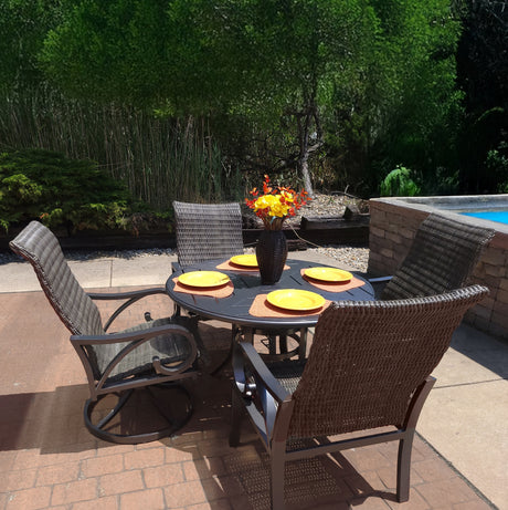 This image shows a brown wicker patio dining set with a round brown table in the center. There are four chairs. On the table, there are four yellow plates on four orange mats. In the center of the plates, there's a vase with yellow and red flowers inside.
