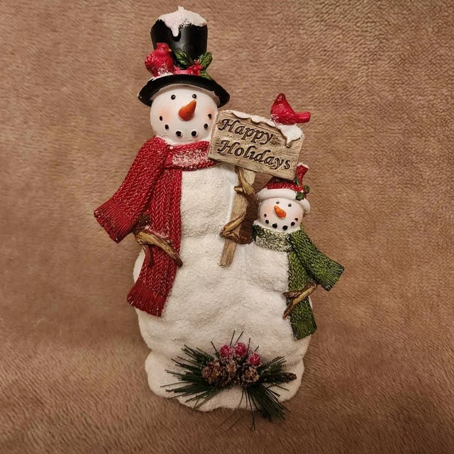 This figurine features a snowman wearing a black top hat and a red scarf and the baby snowman is wearing a red, knit Santa hat and a green scarf. They're holding a sign that reads "Happy Holidays."