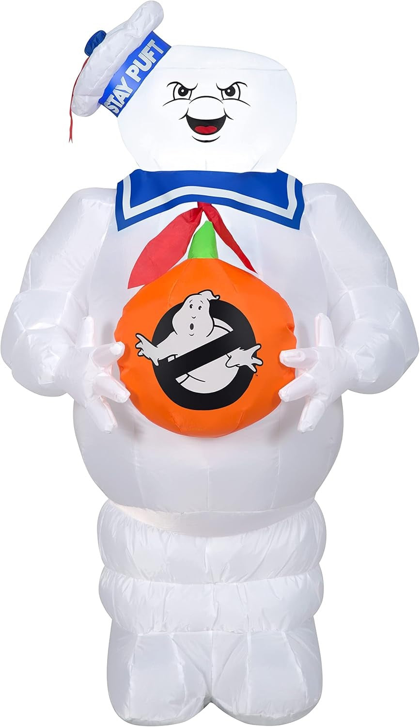 This image shows the Stay Puft Marshmallow Man Halloween Inflatable on a white background. He's wearing a hat that reads "STAY PUFT" and he's holding a pumpkin that has the Ghostbusters logo on it.