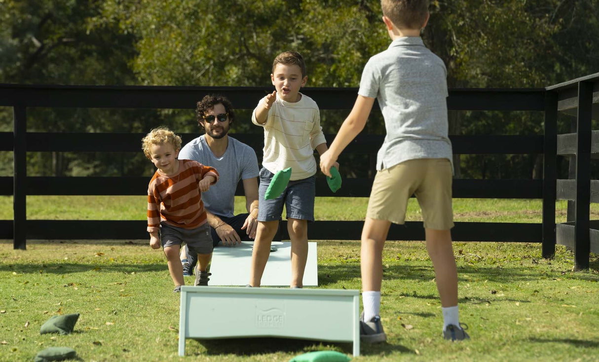 This is a lifestyle image for the cornhole set from Ledge Lounger. This image shows three kids throwing bags while a dad watches from behind.