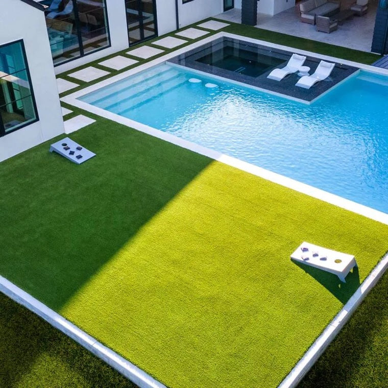 This is a lifestyle image for the cornhole set from Ledge Lounger. It shows two cornhole boards set up in a backyard next to a pool. Inside the pool, there are two pool chairs from Ledge Lounger.