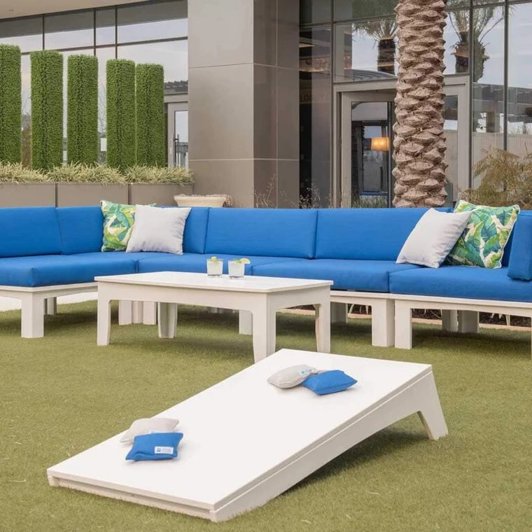 This is a lifestyle image for the cornhole set from Ledge Lounger. This image shows one of the boards on the grass in front of a patio furniture set next to a house.