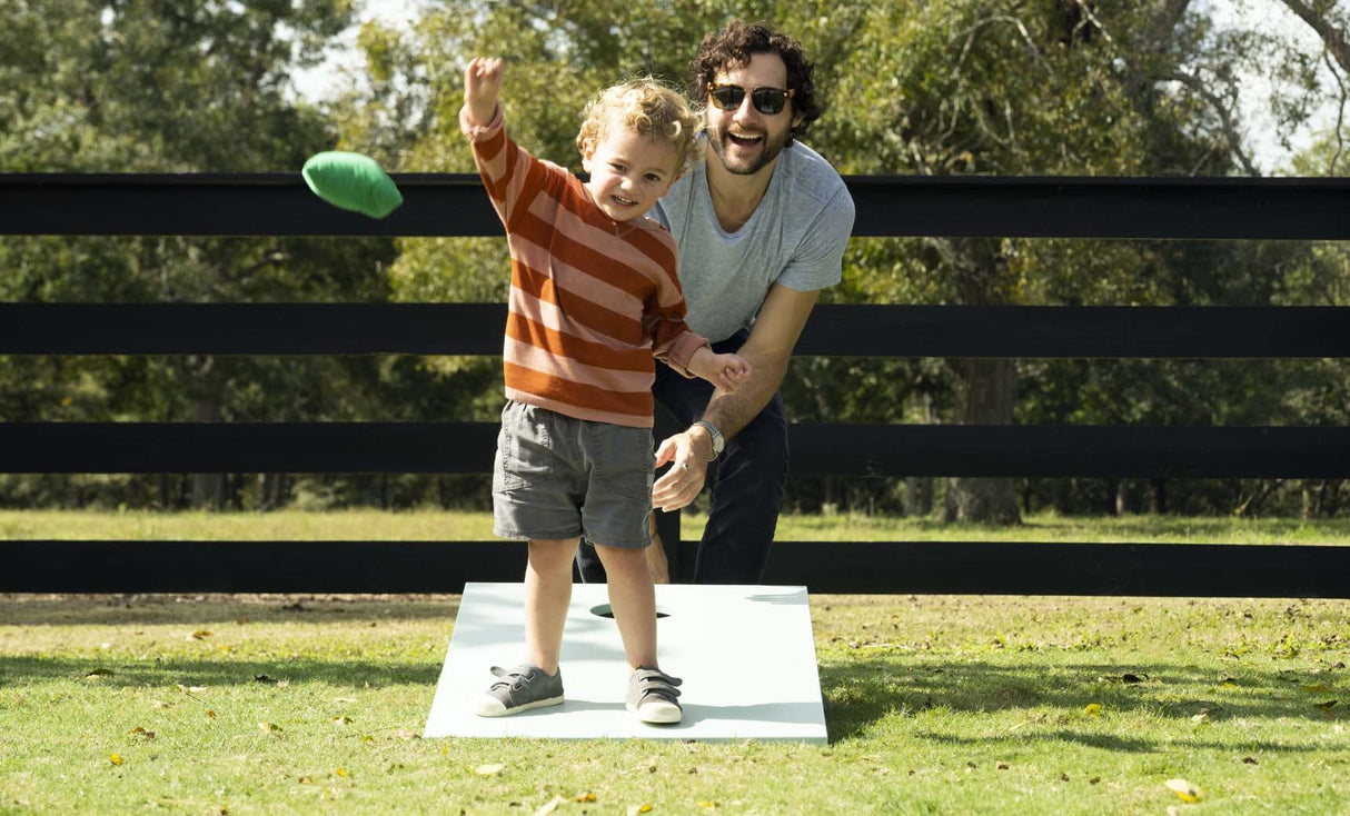 This is a lifestyle image for the cornhole set from Ledge Lounger. This image shows a little boy standing on the cornhole board, throwing a bean bag while his dad cheers him on behind the board.