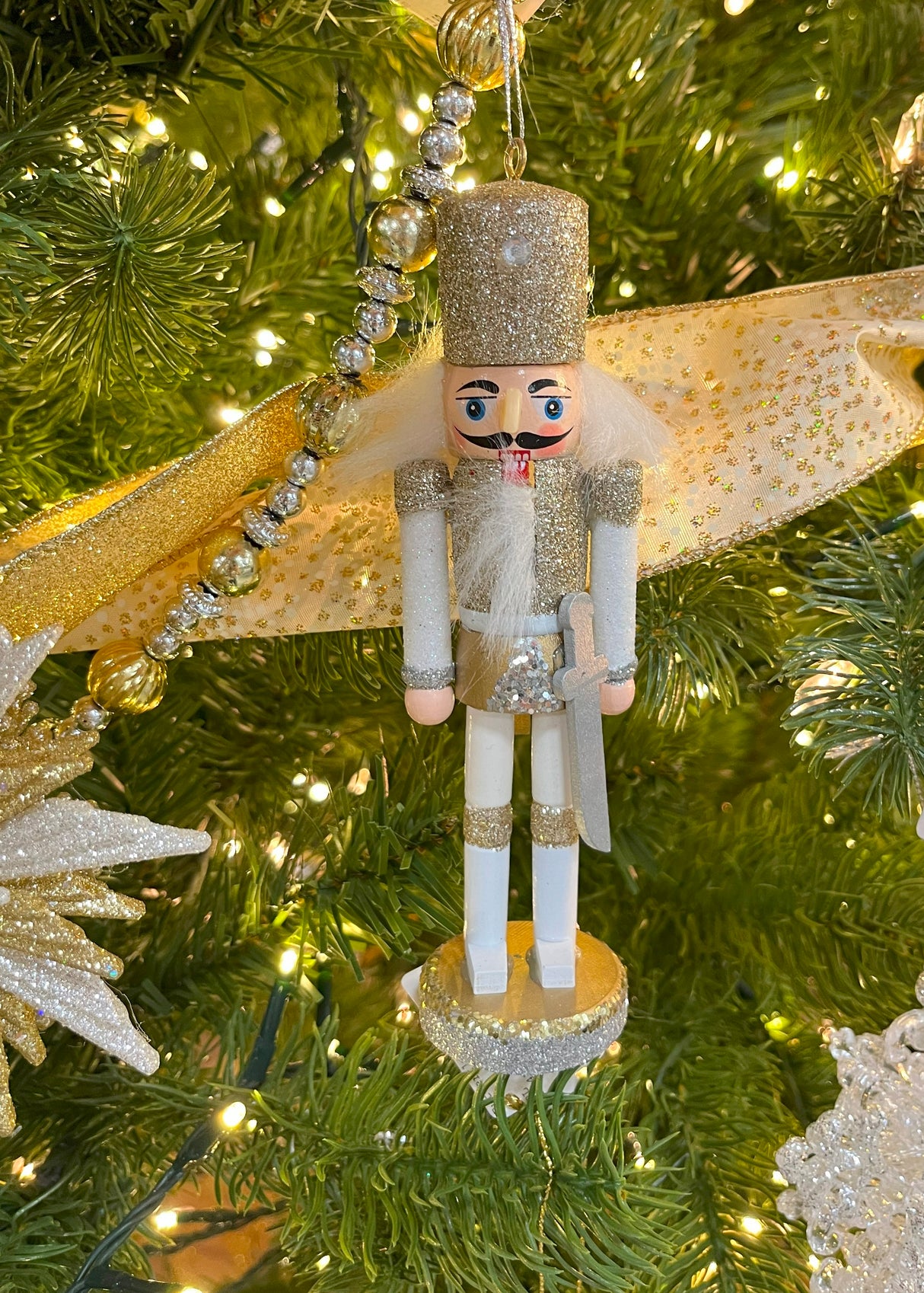 Glittery nutcracker hanging on a Christmas tree with other beaded garlands and ornaments.