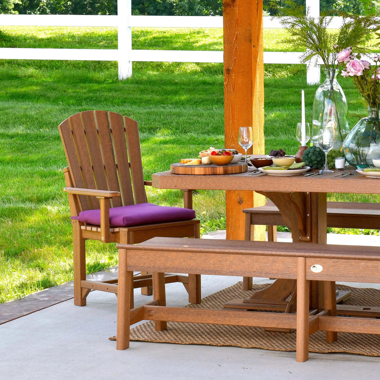 This is a lifestyle image showing the Antique Mahogany colored Adirondack Arm Chair in front of a matching patio dining table and two benches.