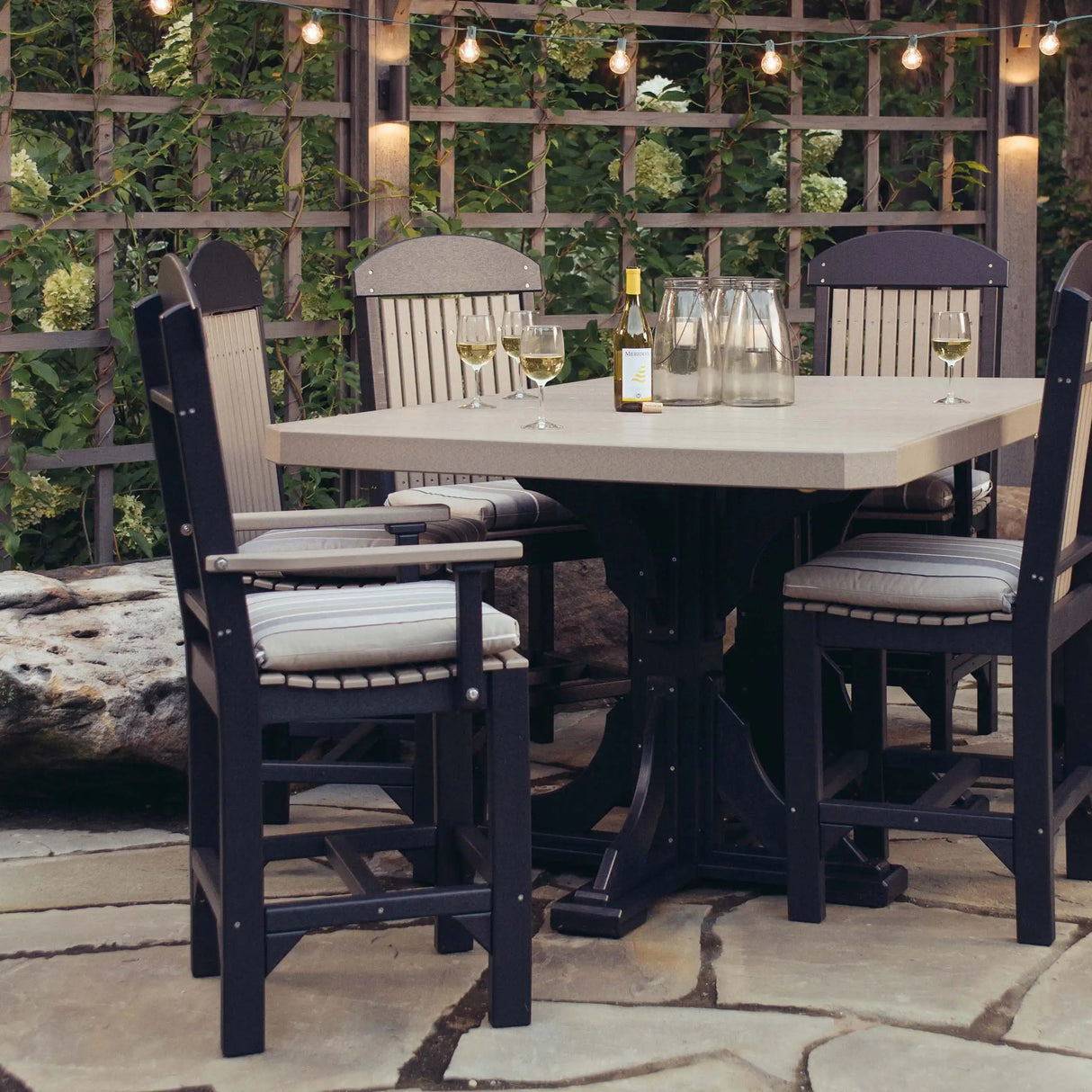 This is a lifestyle image showing Weatherwood and Black colored Arm Chairs around a matching table. It's on a stone patio with a wall lattice behind it. There's a string of Edison lights hanging from the lattice.