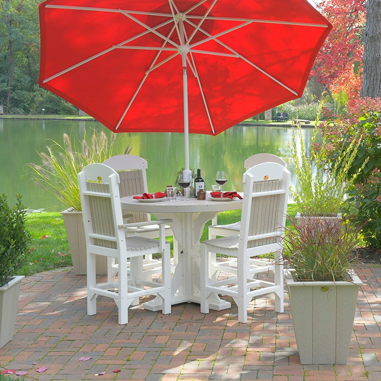 This is a lifestyle image showing four Birch and White colored Classic Arm Chairs around a round, matching patio dining table. They're on a brick patio with a body of water behind them. There's a red umbrella shielding the patio set from the sun.