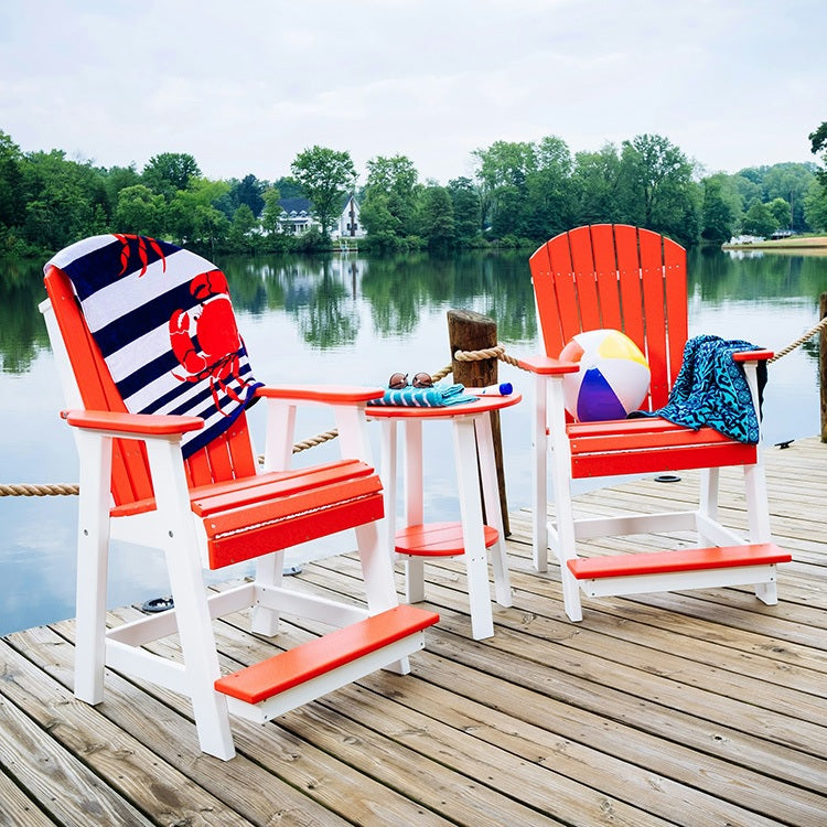 This is a lifestyle image showing two matching Red and White colored Balcony Adirondack chairs with a matching table between them. They're set up on a dock over a body of water.