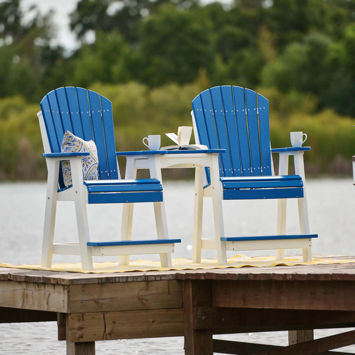 This is a lifestyle image showing two matching Balcony Adirondack Chairs on a pier over a body of water. The chairs and the table are Blue and White.