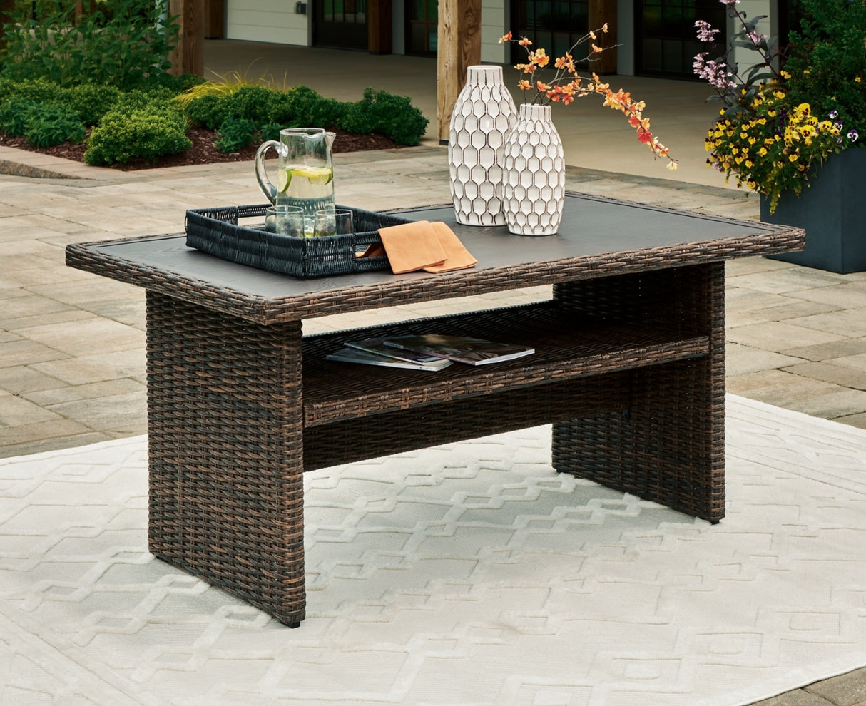 This is a lifestyle image showing this wicker coffee table on an outdoor rug on a stone patio. On top of the table, there are two vases of flowers and a pitcher of lemonade. A few inches underneath the tabletop, there's a shelf. In this image, on the shelf, there are a couple magazines.