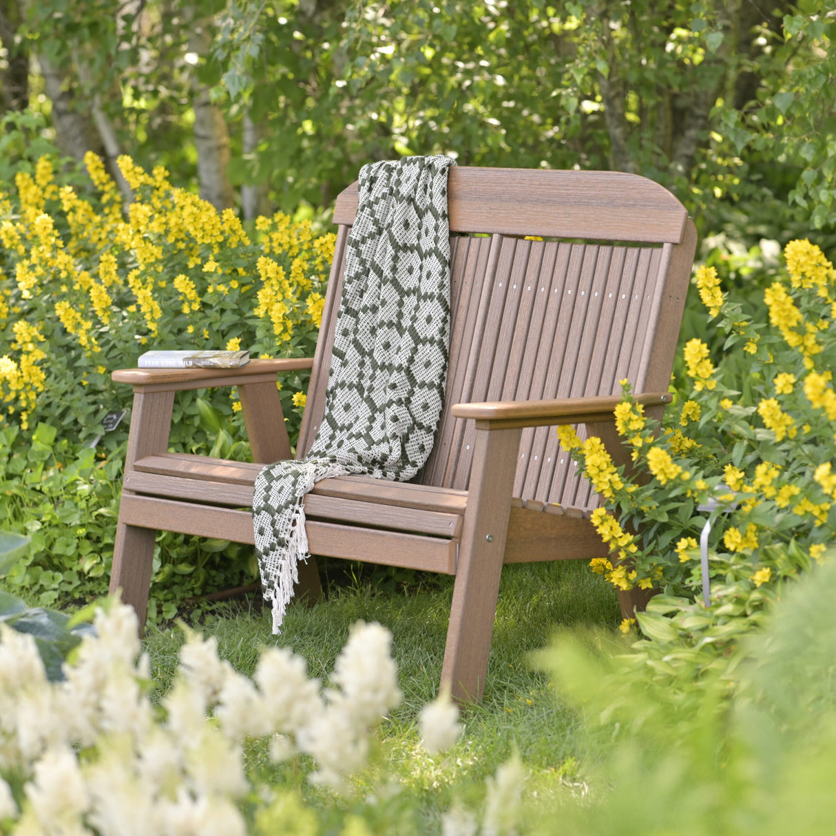 This is the Antique Mahogany colored bench in a grassy yard surrounded by yellow flowers. There's a blanket draped over the side.