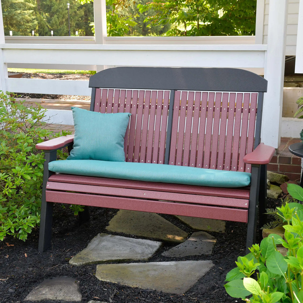 This is a lifestyle image showing the Cherrywood and Black colored bench up against a white fence in a garden. There's a teal colored pillow and seat cushion on it.