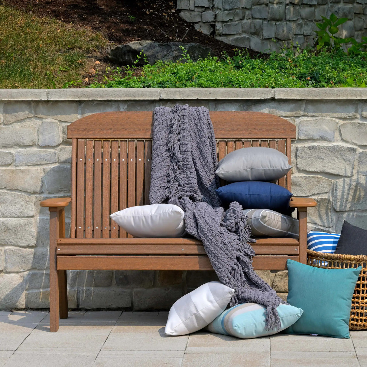 This is the Antique Mahogany colored bench up against a stone fence. There are multiple pillows on the bench and on the ground, and a blanket draped over the side.