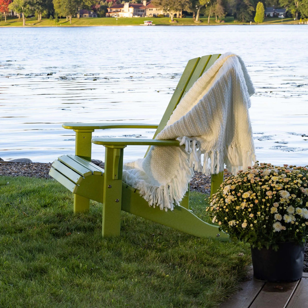 This is a lifestyle image showing the Lime Green Adirondack chair on a grassy patch near a body of water. There's a white blanket draped over the side of the chair.