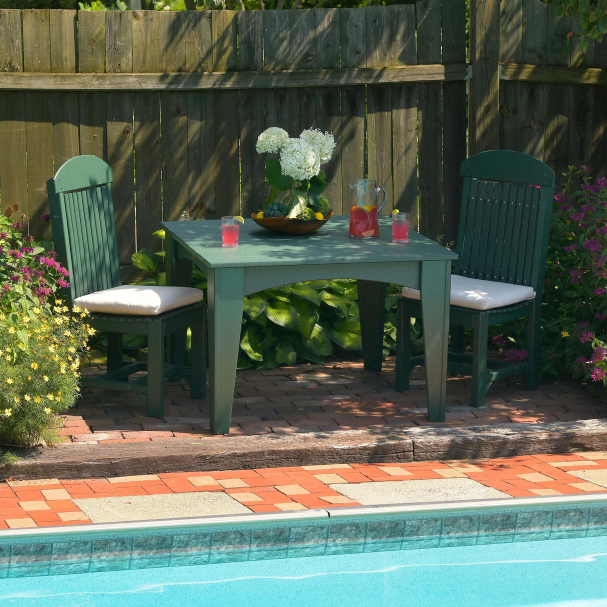 This is a lifestyle image showing two Green Side Chairs with a matching table between them. There's a pitcher of pink lemonade on the table. They're all on a stone patio next to a pool.