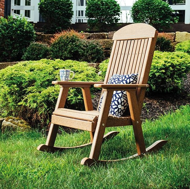 This is a lifestyle image showing an Antique Mahogany colored rocker chair in the grass in front of a house. There's a blue and white pillow on the chair and a blue and white mug on the armrest.
