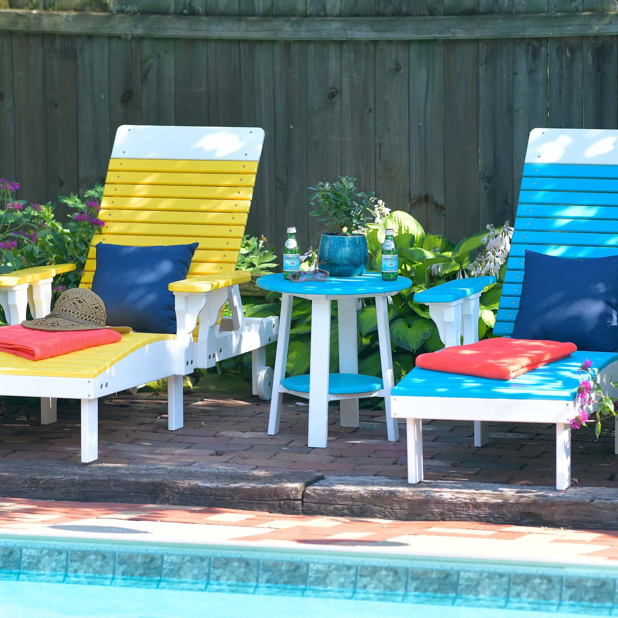 This is the a lifestyle showing two beach chairs near a pool and there's an end table between them. The chair on the left is Yellow and White and has a blue pillow on it. The end table is Aruba Blue and White and has two water bottles on it. The chair on the right is Aruba Blue and White and has a blue pillow on it.