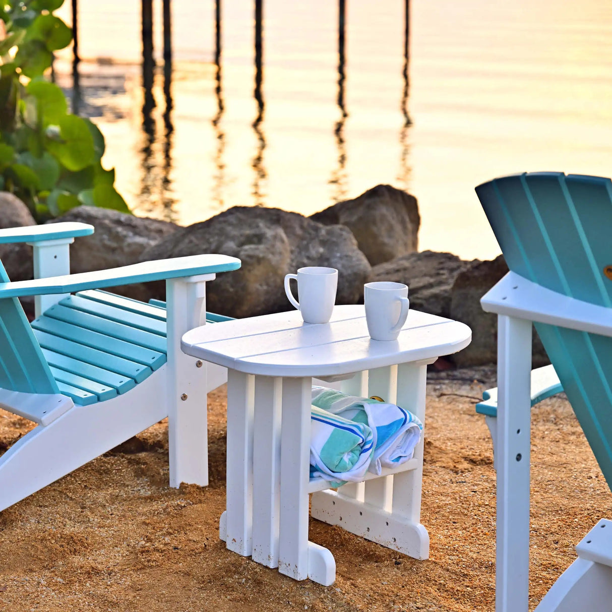 This is a lifestyle image showing the White colored end table in between two Aruba Blue and White colored Adirondack chairs on a beach.
