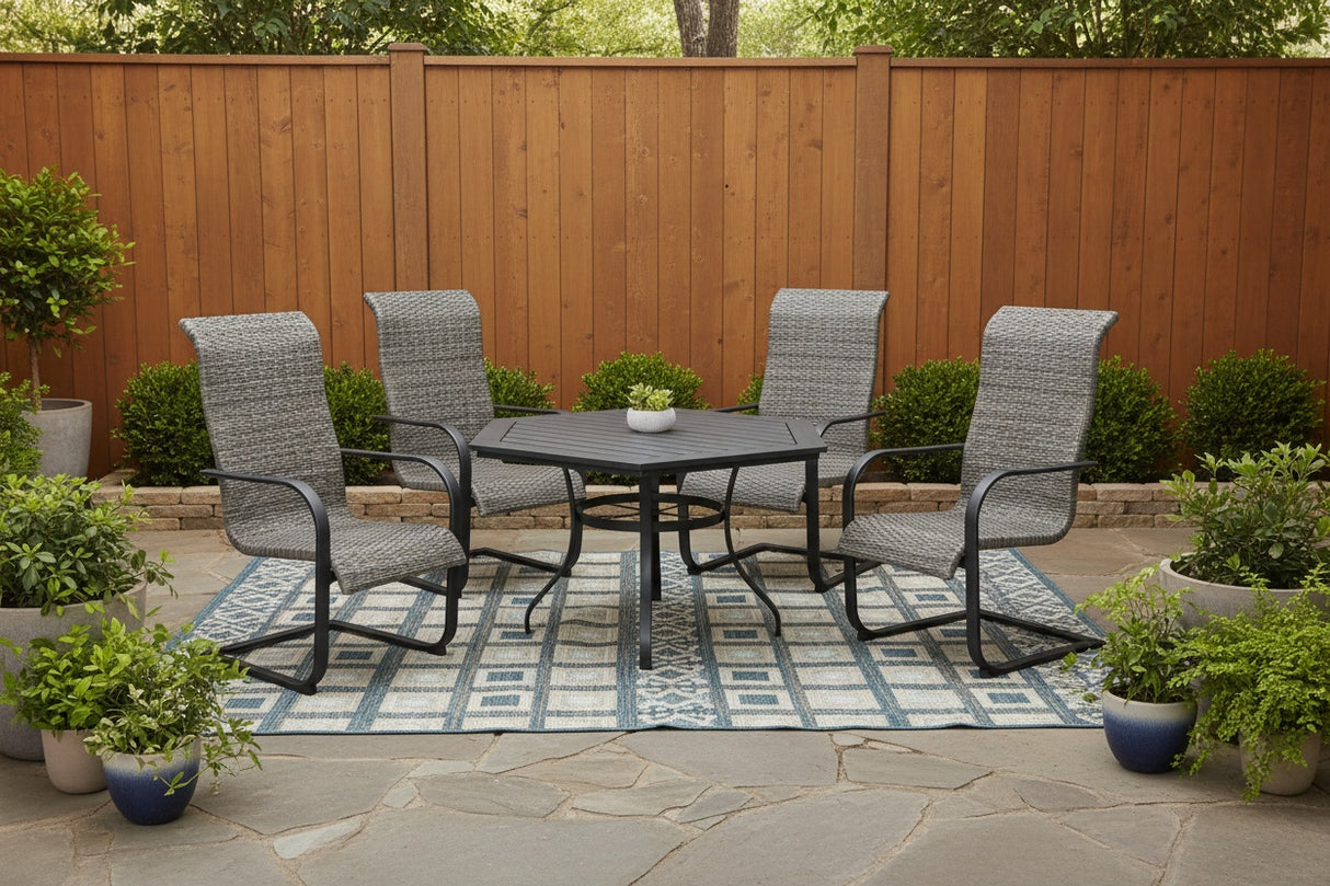 This is a lifestyle image showing these spring wicker chairs and a hexagon dining table on an outdoor rug on a stone patio in a backyard. There are lots of potted plants and a tall, wooden fence.