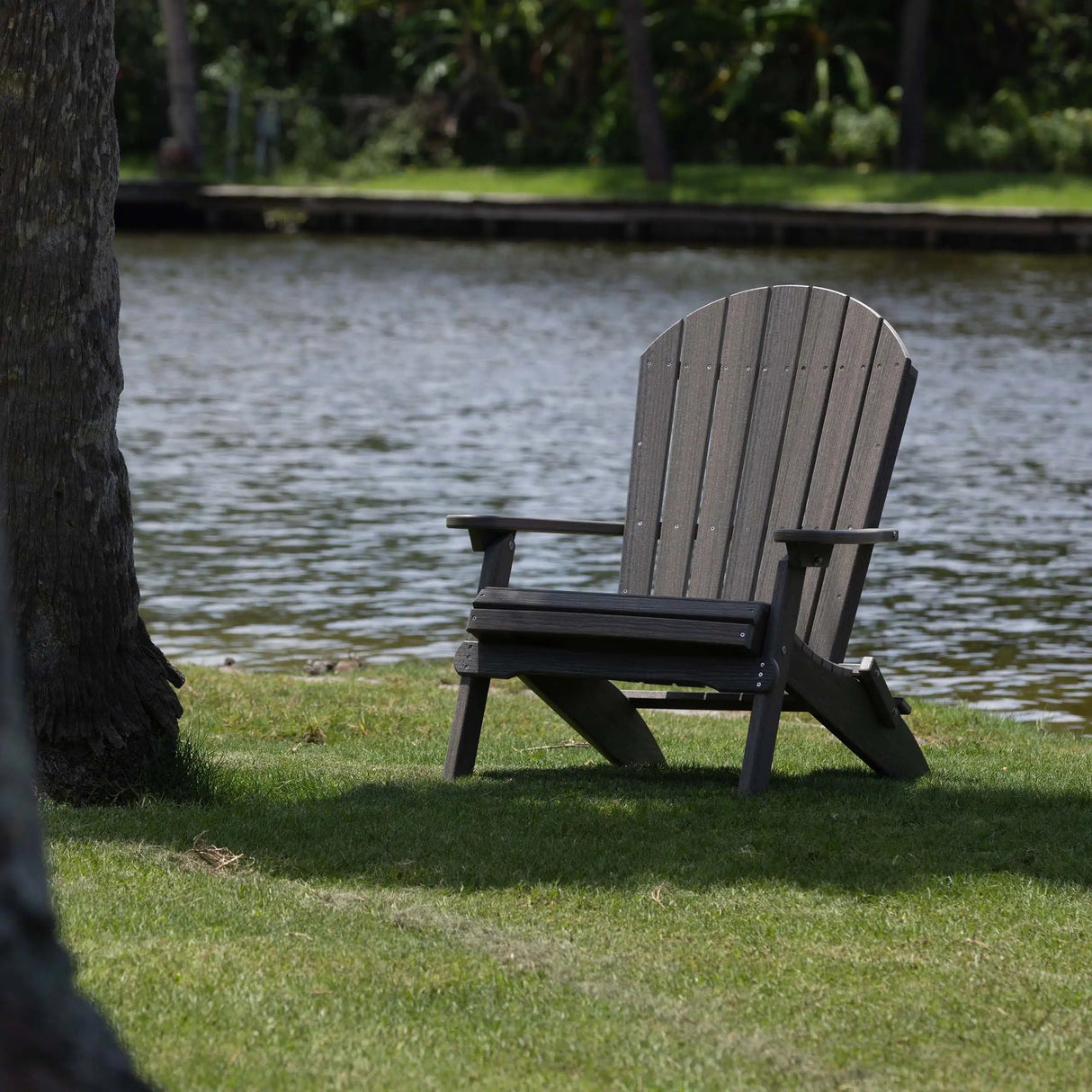 This is the Coastal Gray colored Folding Adirondack chair on a grassy patch under some trees next to a body of water.