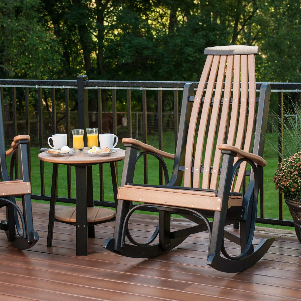 This is a lifestyle image showing two Antique Mahogany and Black colored Grandpa's Rocking Chairs with a matching table between them. They're on a wooden porch in a grassy backyard.