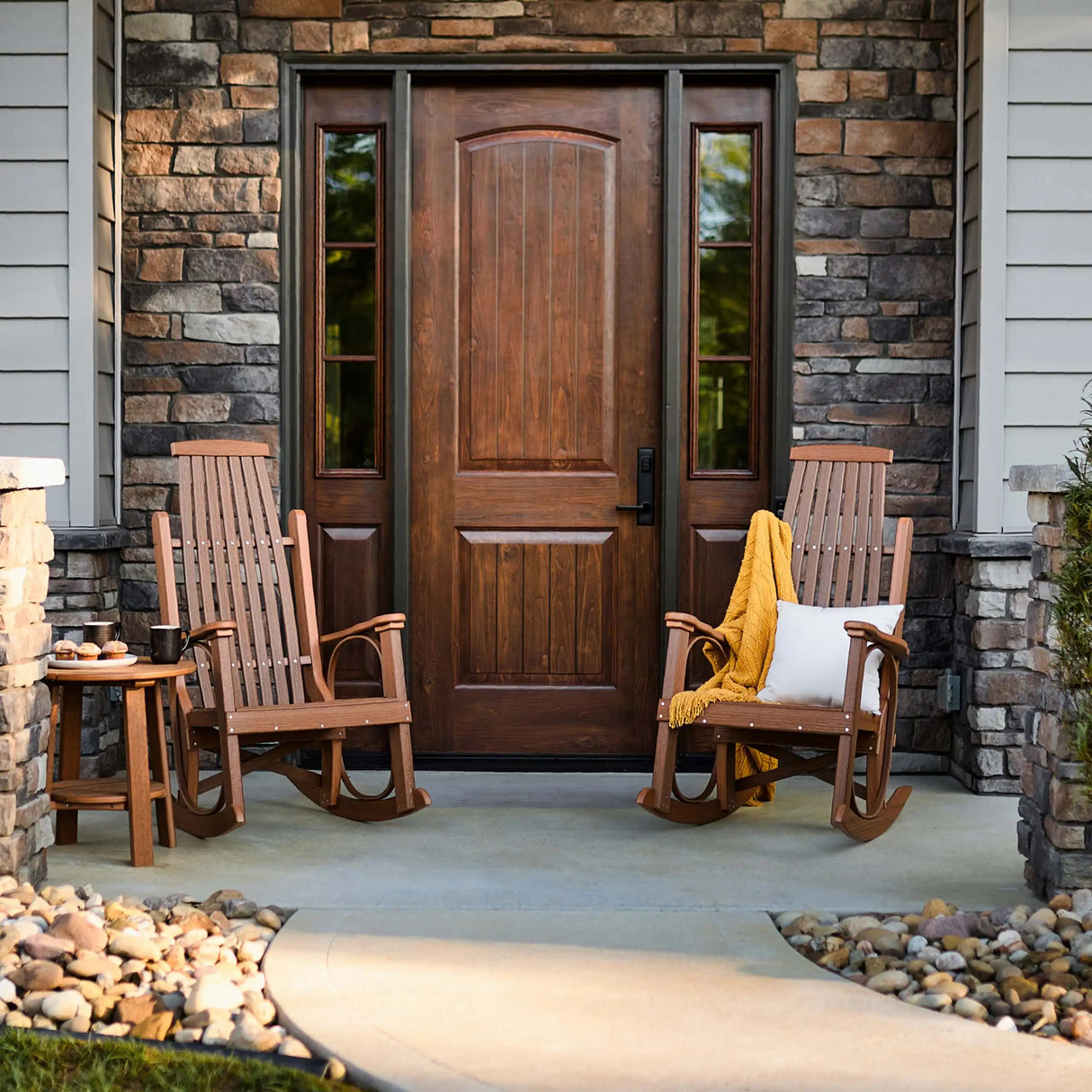 This is a lifestyle image showing two Antique Mahogany colored Grandpa's Rocker Chairs in front of a beautiful wood and brick front door. There's a matching table near the one on the left.