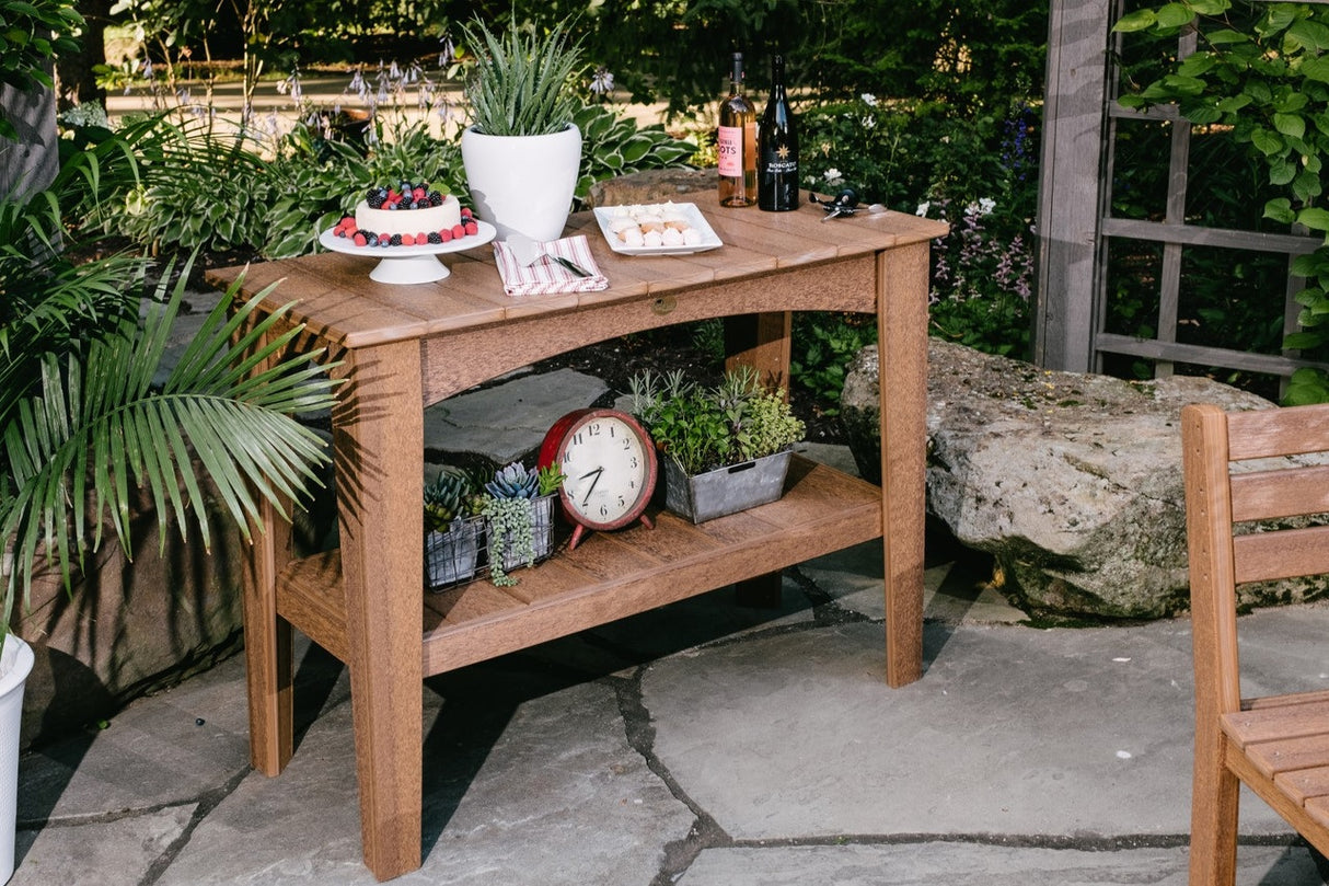 This is a lifestyle image showing the Antique Mahogany colored Island Buffet Table on a stone patio. On top of the table are a cake with berries on it, a vase, a serving spoon, a serving of small cookies and two bottles of wine. The bottom shelf of the table has three different plants and a red clock.