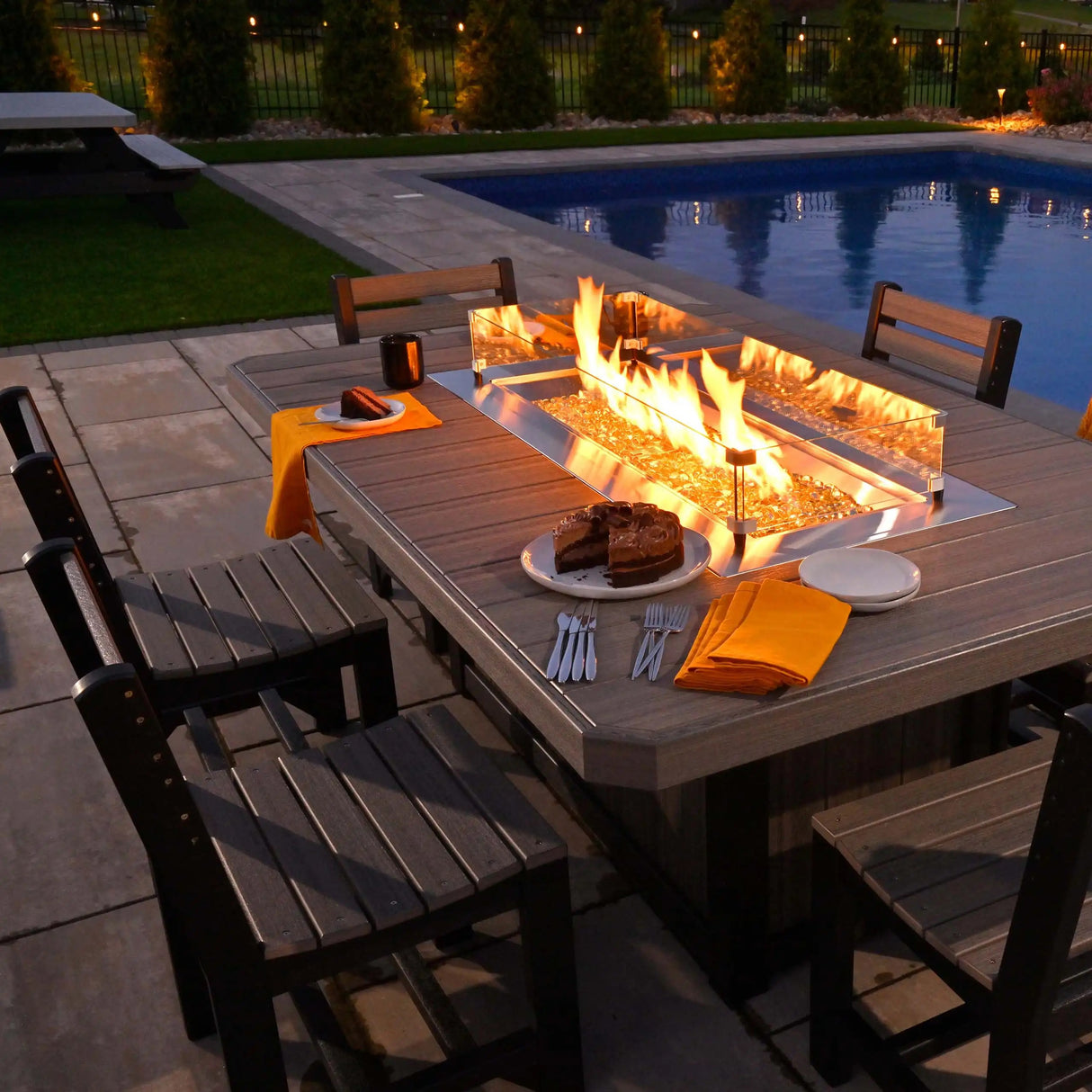 This is a lifestyle image showing six Weatherwood and Black colored Side Chairs around a matching fire pit table on a concrete patio next to a pool. There's a plate with a chocolate cake on the table.