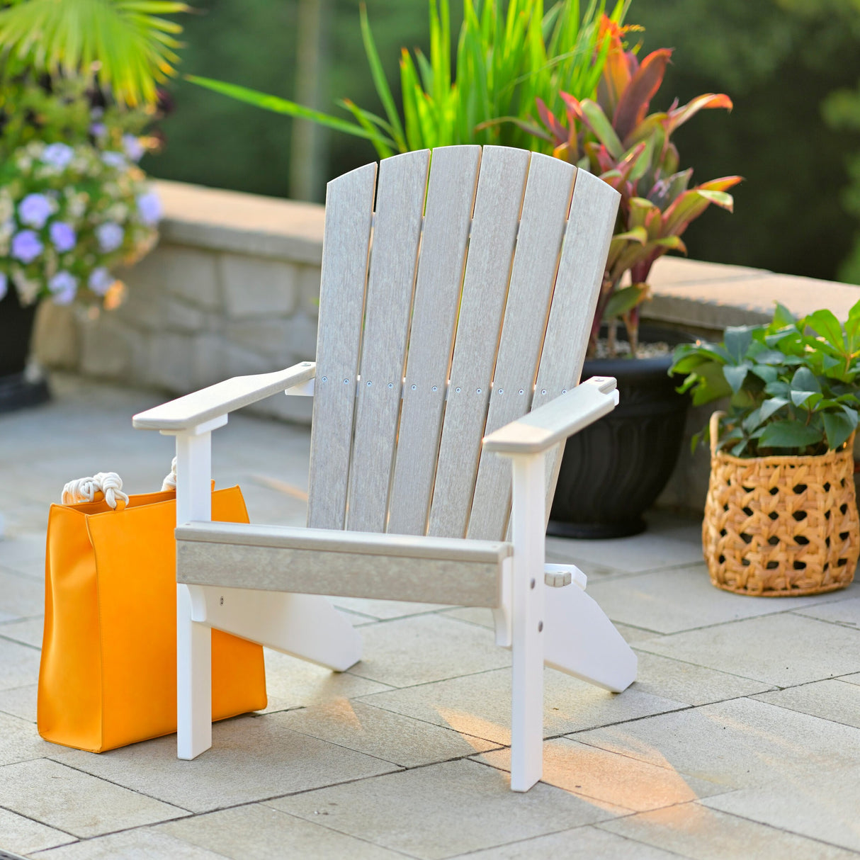 This is a lifestyle image showing the Birch and White colored Lakeside Adirondack Chair on a backyard patio. There's an orange purse on the ground next to the chair and a few potted plants behind it.