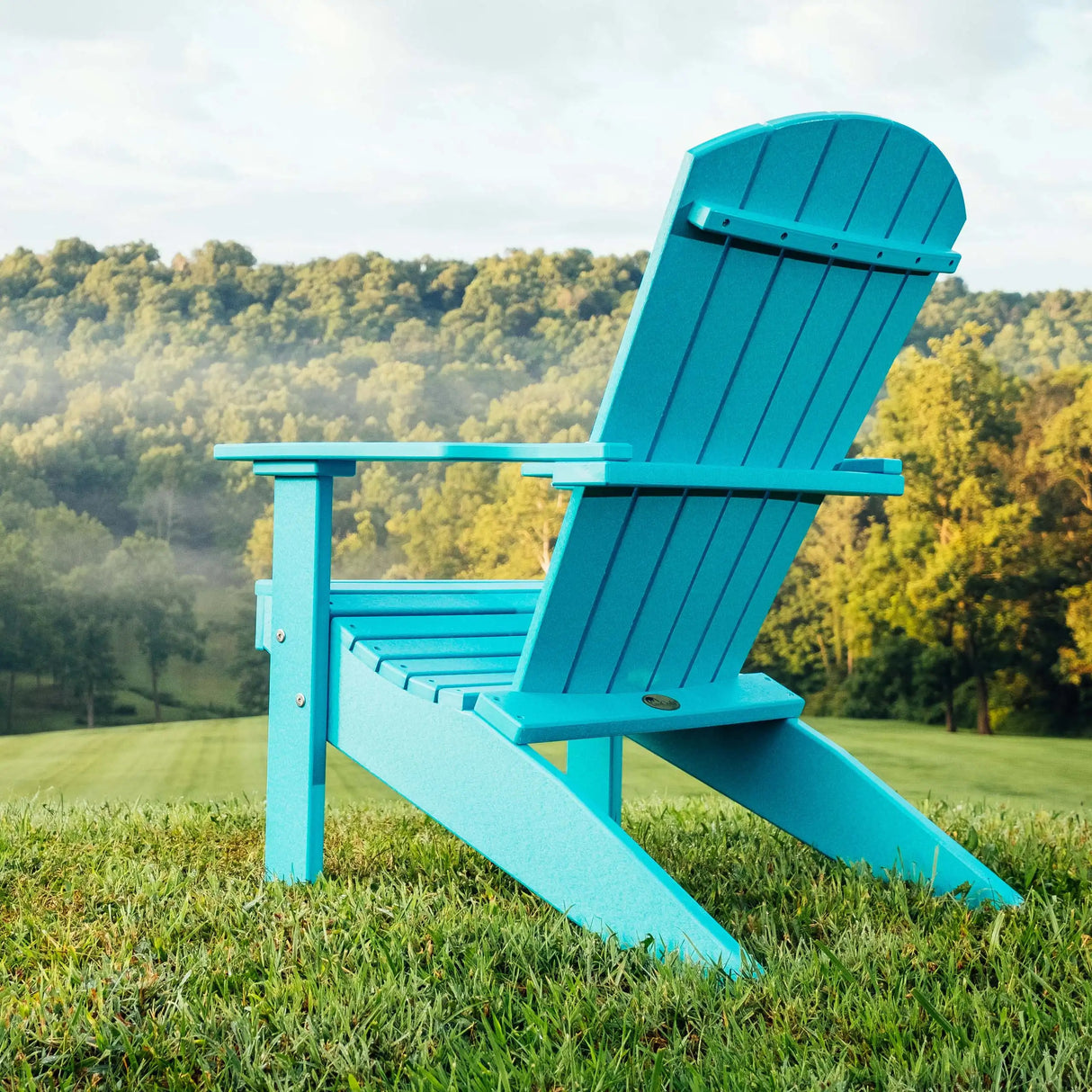 This is a lifestyle image showing the Aruba Blue colored Lakeside Adirondack Chair in a grassy field overlooking a forest.