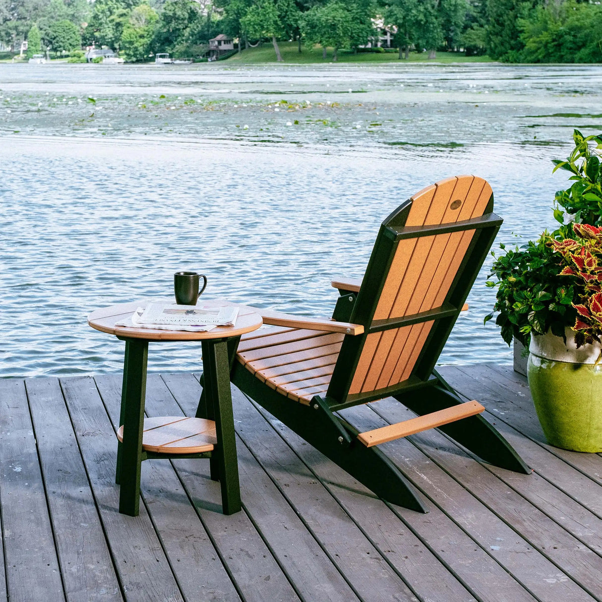 This is a lifestyle image showing the Cedar and Black colored Lakeside Adirondack Chair next to a matching table on a pier overlooking a body of water.