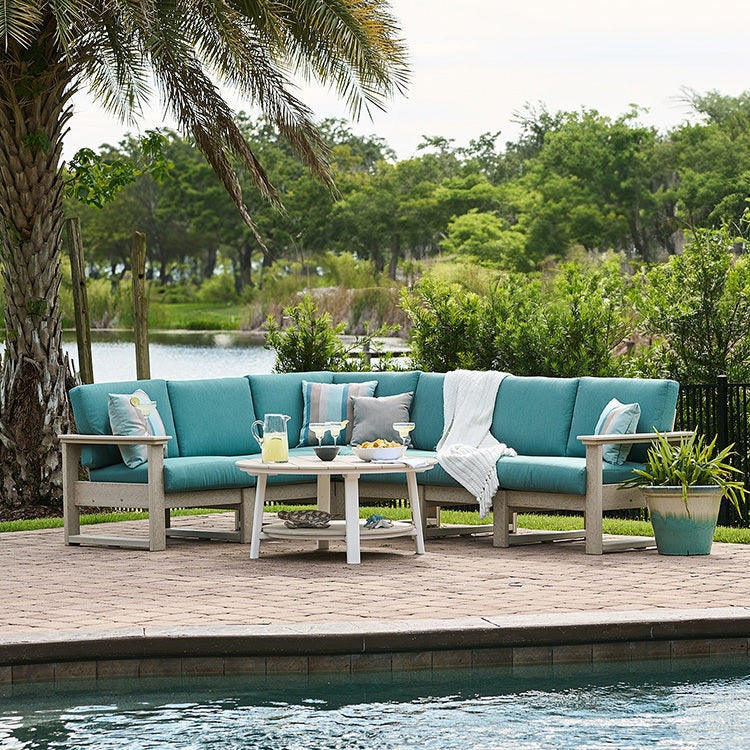 This is a lifestyle image showing an outdoor sectional on a brick patio in front of a pool. The frames are Birch and the cushions are Cast Breeze colored.