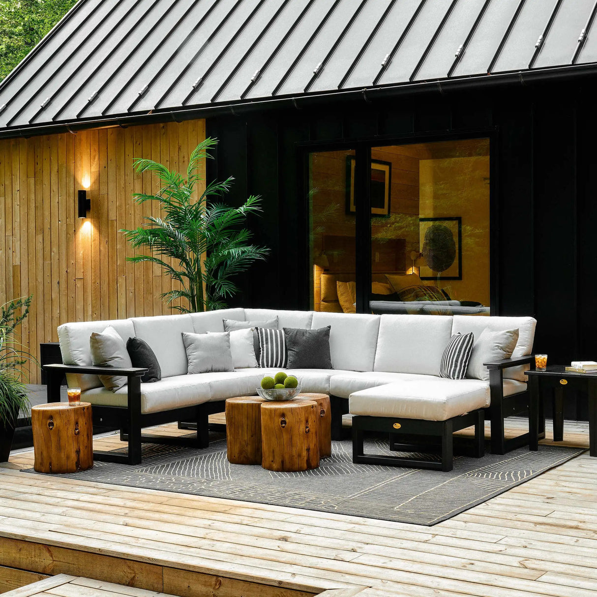 This is a lifestyle image showing a deep seating sectional sofa on a wooden porch outside of a house. The frames are Black and the cushions are Canvas colored.