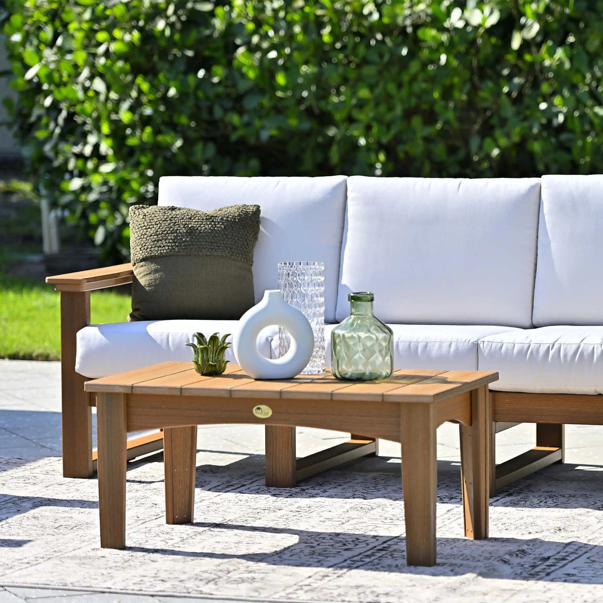 This is a lifestyle image showing a sofa in front of a matching table on a cement patio in a grassy backyard. The frames are Antique Mahogany and the cushions are Canvas colored.