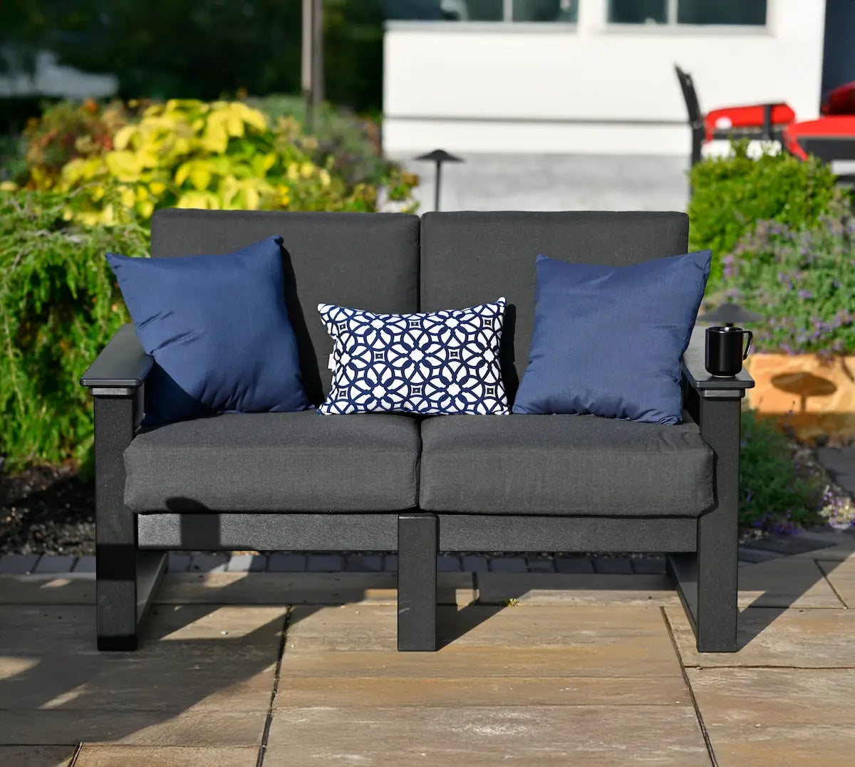 This is a lifestyle image showing the deep seating love seat on a stone patio in a backyard with lots of plants. The frames are black and the cushions are spectrum carbon. There are two navy blue throw pillows and one white and navy blue pillow with a unique, floral-esque design. There's a black mug on one of the armrests.