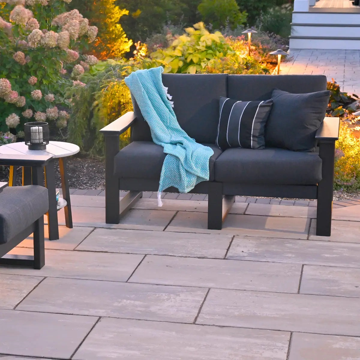 This is a lifestyle image showing the deep seating loveseat on a stone patio in a backyard with lots of plants. Off to the side of the image, you can see a small, circular table and a deep seating chair. The frames are black and the cushions are spectrum carbon.