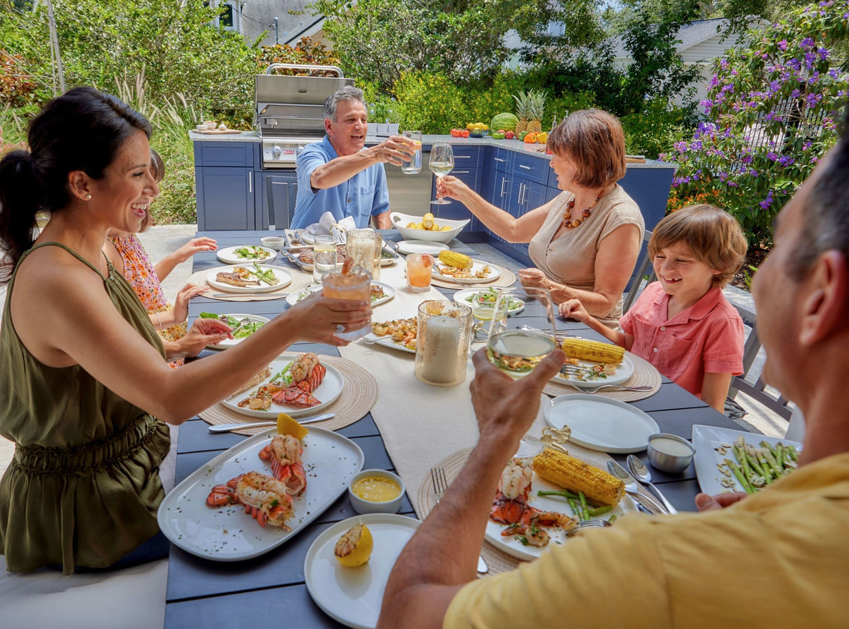A family of 6 are gathered together eating seafood and corn on the cob. They're all laughing and toasting their drinks. In the background is a DuraBULL outdoor kitchen.